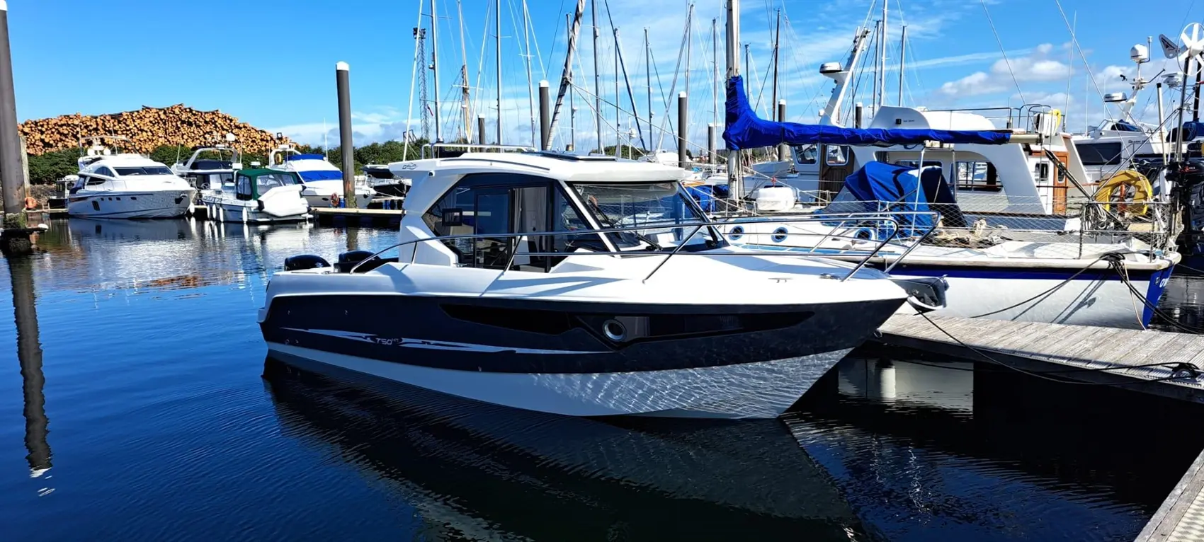 A sleek motorboat docked at a marina, surrounded by various boats and a pile of logs in the background.